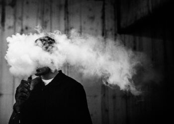A dark and high contrast image of a young adult male has a cloud of nicotine vapor "smoke" pouring from his mouth.  Vape smoking, or "vaping" is growing in popularity, as well as falling under stricter state and governmental regulations.  Horizontal image with copy space.