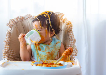 Adorable little toddler girl or infant baby drink water after eat delicious spaghetti food on chair. Funny cute infant girl get thirsty. Lovely mix race daughter get dirty. Kid hold spoon upside down