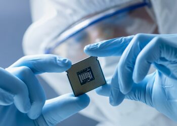 In Ultra Modern Electronic Manufacturing Factory Design Engineer in Sterile Coverall Holds Microchip with Gloves and Examines it.