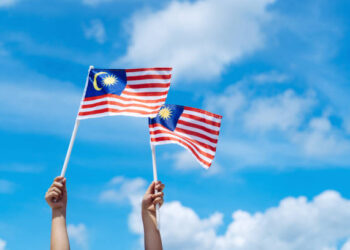 Hand happily waving Malaysia flag against blue sky. Celebrate Independence Day & Merdeka day.