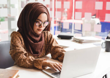 Young asian muslim business woman in smart casual wear working with laptop computer while sitting in the creative cafe.